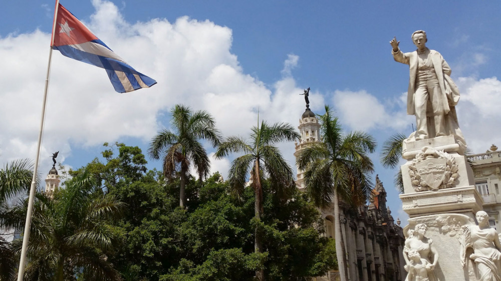Statue von Jos Marti im Parque Central in Havanna mit Hotel Inglaterra, Theater Alicia Alonso und das Capitolio von Havanna im Hintergrund. Blauer Himmel mit weien Wolken und die kubanische Flagge ()